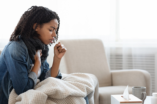 Risk Of Coronavirus. Sick Black Woman Coughing Hard At Home, Sitting On Couch Wearing Scarf And Covered With Blanket, Side View With Copy Space