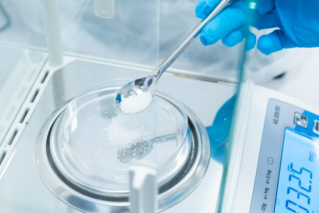 Close up scientist hands in rubber gloves use metal spatula for weighing white powder of the substance using digital balances. Preparation to clinical or pharmaceutical analysis.
