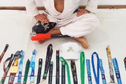 Uma pessoa em um gi branco de artes marciais senta-se no chão com uma variedade de faixas coloridas, medalhas e um boné branco entre os itens, exibindo sua dedicação. Com uma resolução inabalável refletida em sua perna protética, talvez eles encontrem relaxamento por meio de práticas conscientes como meditação com cannabis.