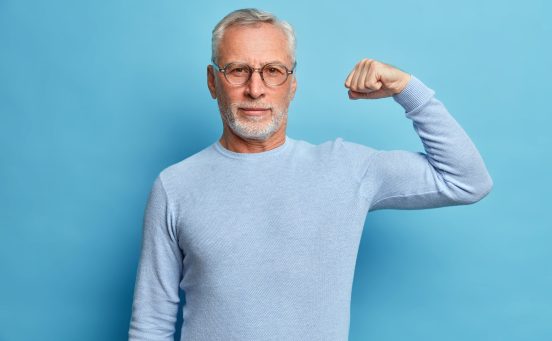 senior-bearded-man-shows-muscles-after-practising-bodybuilding-wears-transparent-glasses-basic-jumper-poses-against-blue-studio-wall-scaled-1.jpg