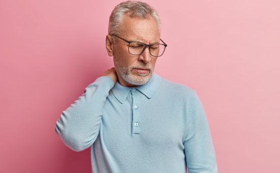 Image of dissatisfied elderly man touches neck, suffers from pain, closes eyes from displeasure, wears optical glasses, casual jumper, poses against pink background, feels unwell and overworked